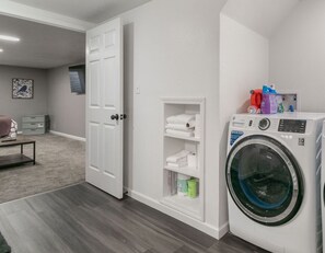 A laundry room with a front-loading washing machine, a shelf with folded towels, and a door leading to another room. - Bluebird House - Extended Stay (Great Falls)