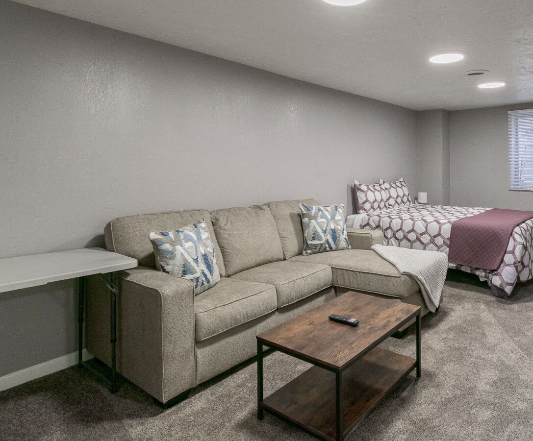 A modern, compact living space with a sectional sofa, a small coffee table, and a bed with patterned bedding.