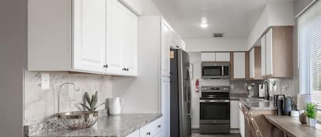 Cocina moderna con armarios blancos, encimera de granito, refrigerador de acero inoxidable, microondas y horno empotrado. Hay sillas de madera y una pequeña barra con fregadero y encimera.