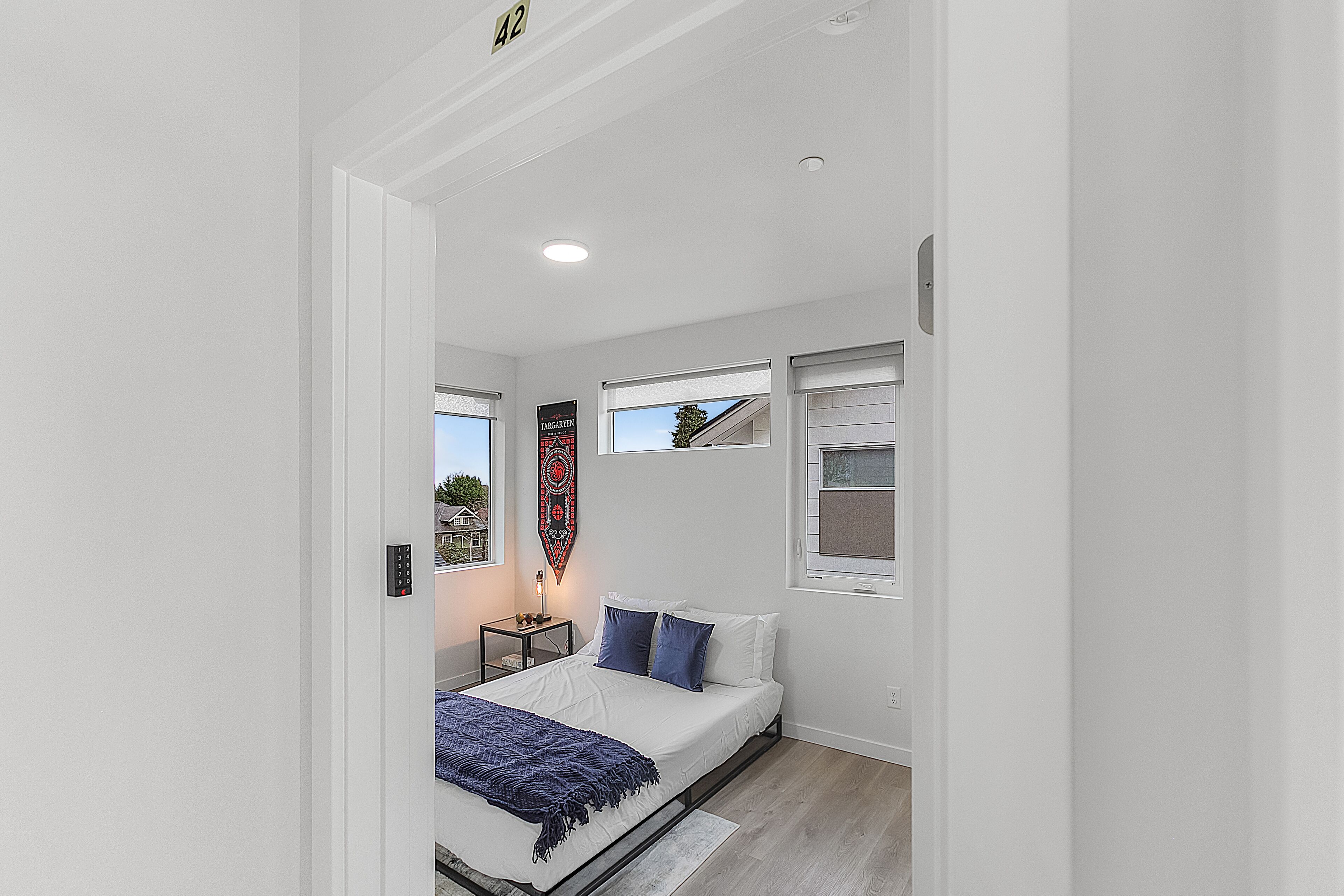 Une chambre moderne avec un lit, deux fenêtres, une table de chevet et un luminaire fixé au mur.