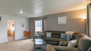 A living room with a sofa, a coffee table, and a chair. There is a window with curtains, a lamp, and a painting on the wall. - Buffalo Jump Retreat - Extended Stay (Great Falls)