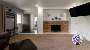 A living room with a fireplace, a television, and a door leading to another room. - Buffalo Jump Retreat - Extended Stay (Great Falls)