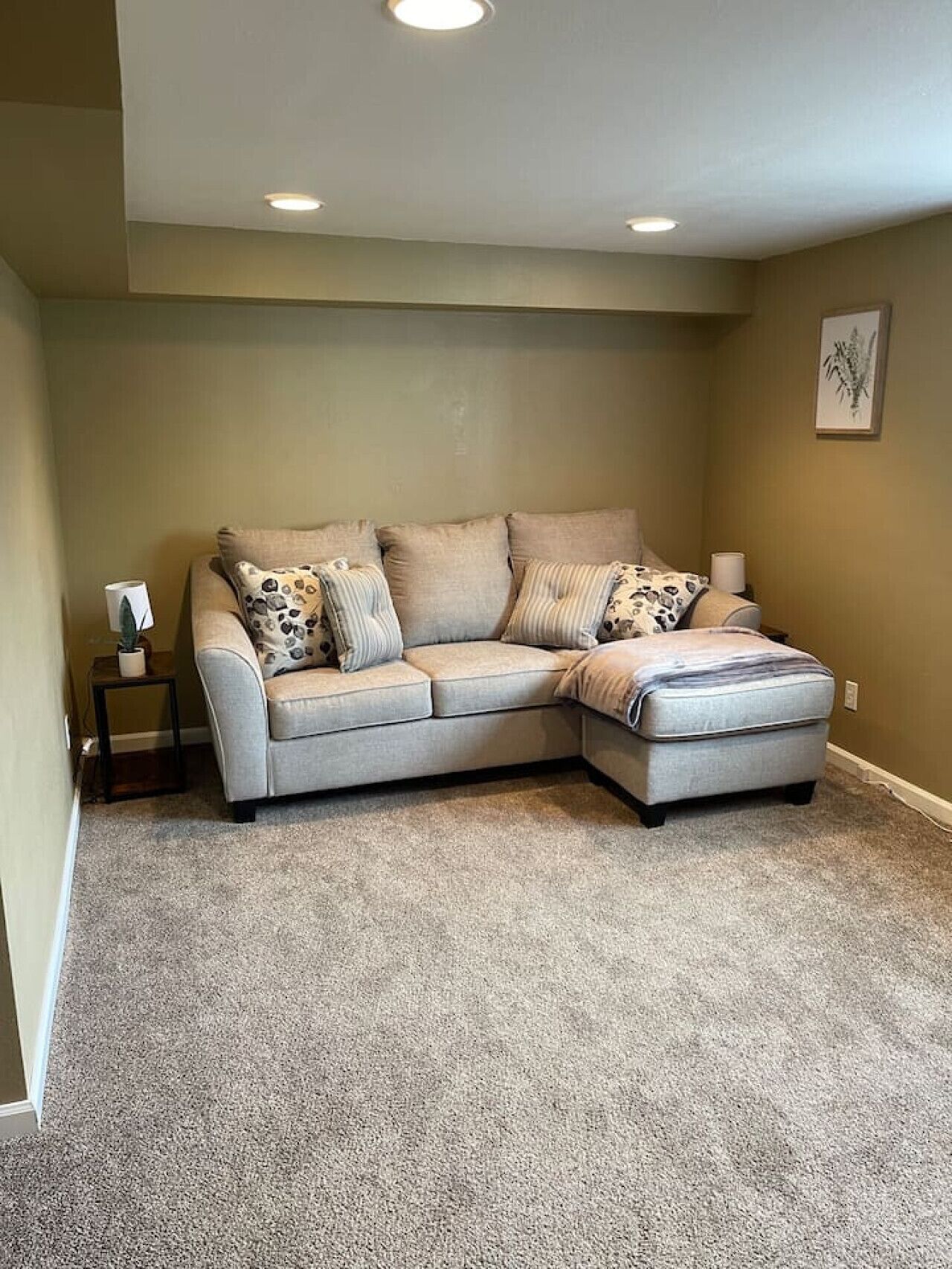 A corner living room with a beige sectional sofa, a small side table, and a carpeted floor.