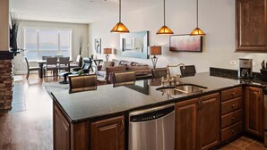 A spacious kitchen with a central island, wooden cabinets, and a dishwasher. There is a dining area with chairs and a table, and a large window offering a view. - Elevated Waterfront | Pool, Beach & Near Downtown (Sandpoint)