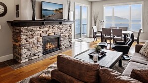 A living room with a stone fireplace, a television, and a dining area with a view of the sea. - Elevated Waterfront | Pool, Beach & Near Downtown (Sandpoint)