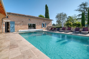 A swimming pool with lounge chairs, a brick building with a door, and a tall tree in the background.