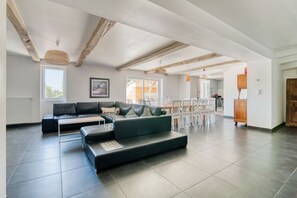 A spacious living area with a sectional sofa, dining table, and chairs, wooden beams on the ceiling, and large windows.