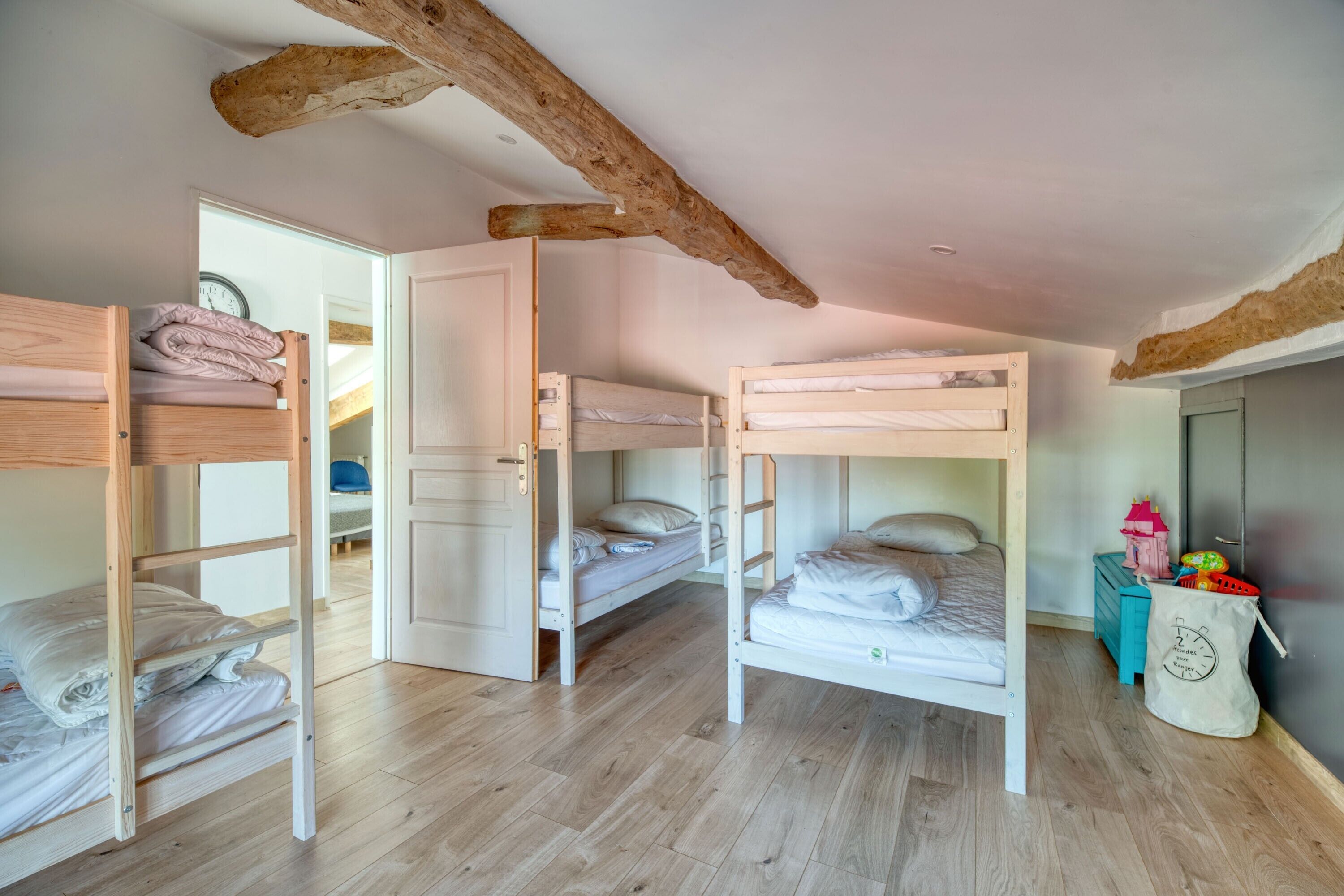 Een kamer met drie stapelbedden, houten balken in het plafond, een deur naar een andere kamer en een kleine tafel met speelgoed.