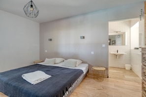 Quarto moderno com uma cama grande, mesas de cabeceira, um banquinho redondo e um banheiro visível através de uma porta aberta.
