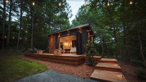 Uma cabana moderna com varanda, cercada por árvores, e um caminho de cascalho que a leva até ela.