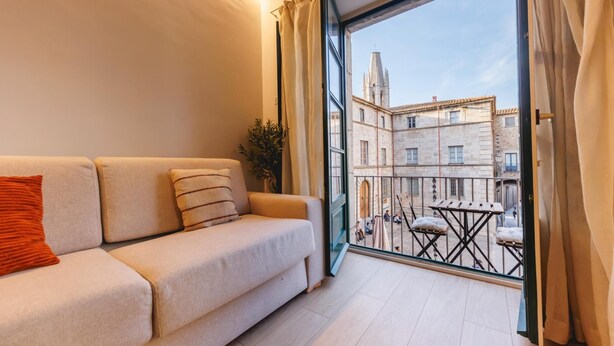 A modern living room with a sofa, a potted plant, and a balcony with a view of historic buildings. - Bravissimo Catedral, with monumental views (Girona)