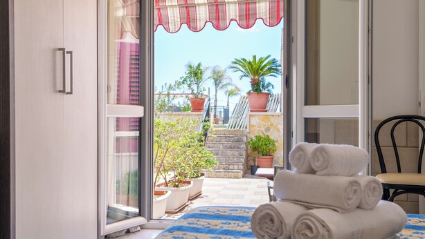A room with a bed, a chair, a table with towels, and a view of the outside balcony with potted plants. - HOTEL LE MACINE (Santa Cesarea Terme)