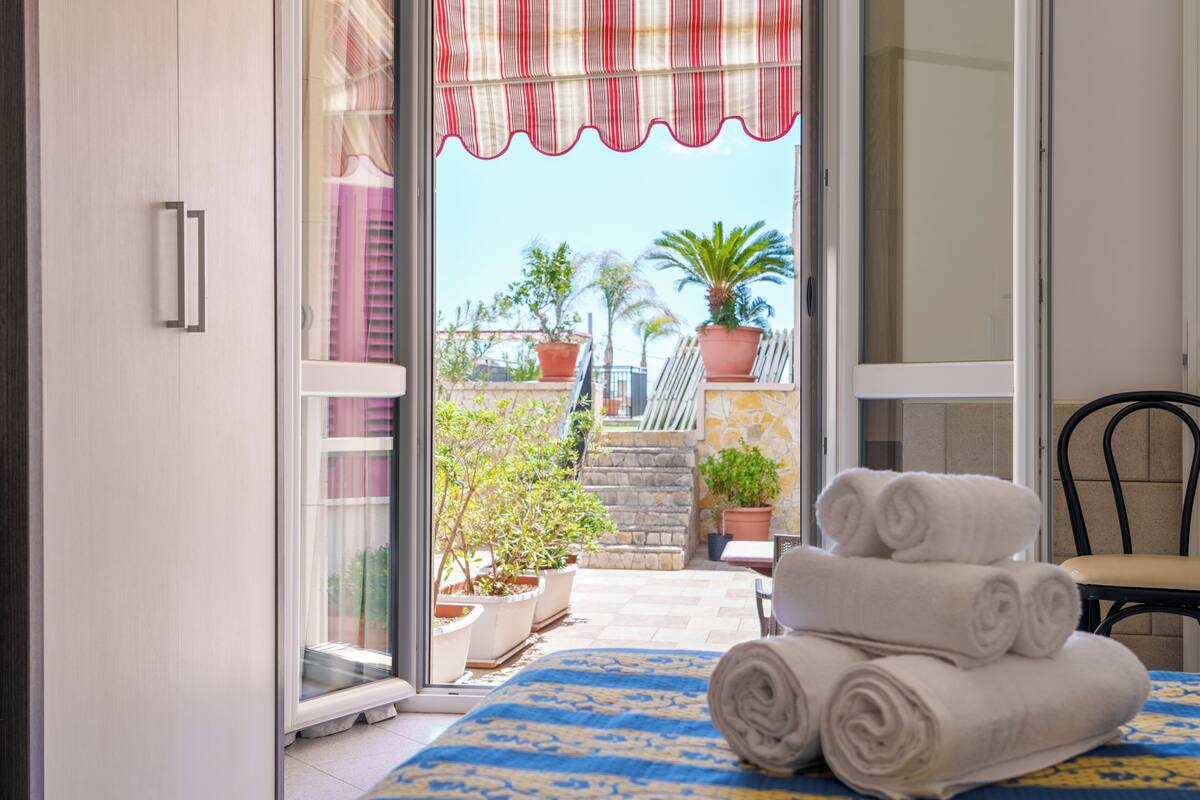 Une chambre avec un lit, une chaise, une table avec des serviettes et une vue sur le balcon extérieur agrémenté de plantes en pot.