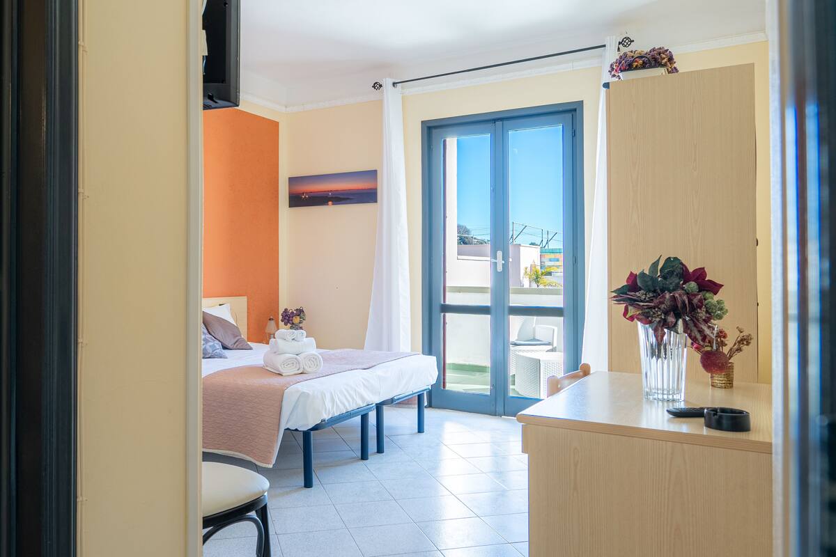 Une chambre d’hôtel avec un lit, un bureau, une chaise, un vase de fleurs et une vue sur l’extérieur, visible à travers une porte vitrée.
