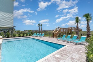 A rectangular swimming pool with blue water, surrounded by a brick patio. There are several blue lounge chairs and two palm trees.
