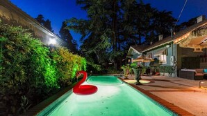 A swimming pool with a red swan float, surrounded by greenery and a house with outdoor lighting. - Boho Vibe House with Pool near Hollywood (Los Angeles)