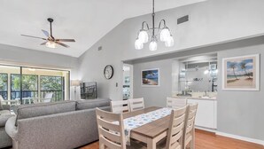 A modern living space with a dining area, gray sofa, wooden dining table, and kitchen in the background.