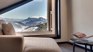 Quarto com uma grande janela oferecendo vista para as montanhas, um sofá aconchegante e uma mesa de leitura.