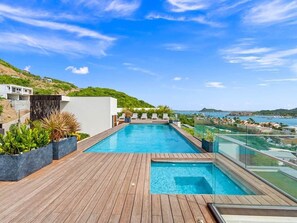Ein Pool auf dem Dach mit Holzterrasse und Pflanzen, mit Blick auf eine Küstenlandschaft mit Hügeln und blauem Himmel.