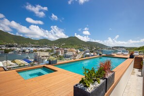 Piscina en la azotea con cubierta de madera, rodeada de plantas en macetas, con vistas a un pueblo costero y colinas.