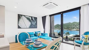 A modern dining area with a wooden table set for four, blue chairs, and a view of a coastal town through large windows. - A136 - Amazing Spacious Apartment Lagoon View with Balcony (Sint Maarten)