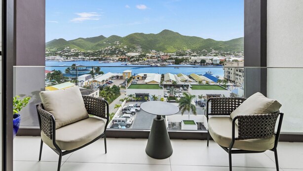 A balcony with two wicker chairs and a small table, overlooking a marina and a coastal town with green hills in the background. - A136 - Amazing Spacious Apartment Lagoon View with Balcony (Sint Maarten)