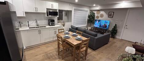 A modern kitchen with white cabinets, a built-in microwave, and a sink. There is a dining area with a wooden table and chairs, and a gray sofa in the living room. A television is mounted on the wall, and a small Christmas tree is visible.