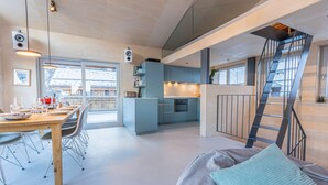 Una cocina moderna con una isla central, un comedor con una mesa y sillas de madera, y una escalera que sube a un nivel superior.