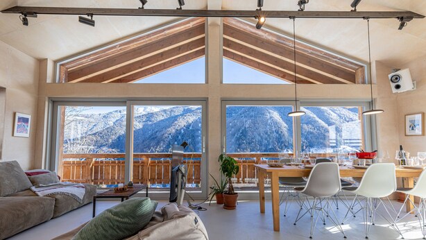 Amplia sala de estar con un gran ventanal con vistas a montañas nevadas, techo de madera y una mesa de comedor con sillas.