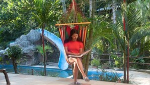 A person sitting in a hammock chair by a pool, with a water slide and tropical vegetation in the background.