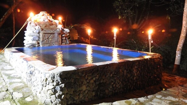A stone pool with a stone wall and a small waterfall feature, surrounded by trees and lit by multiple candles.