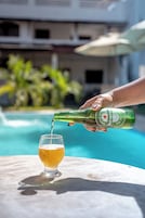 Una mano sirviendo cerveza en un vaso junto a una mesa al borde de la piscina, con vista clara a un edificio al fondo.