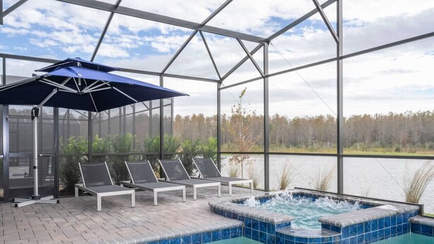A covered outdoor swimming pool with lounge chairs and a hot tub, surrounded by a glass enclosure with a view of a forest and lake.