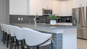 A modern kitchen with white cabinets, a stainless steel refrigerator, a microwave, and a large island with bar stools.