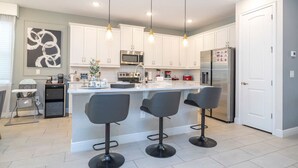 A modern kitchen with white cabinets, a central island with bar stools, stainless steel appliances, and pendant lighting. - House Resort at Encore Reunion - Disney (Kissimmee)