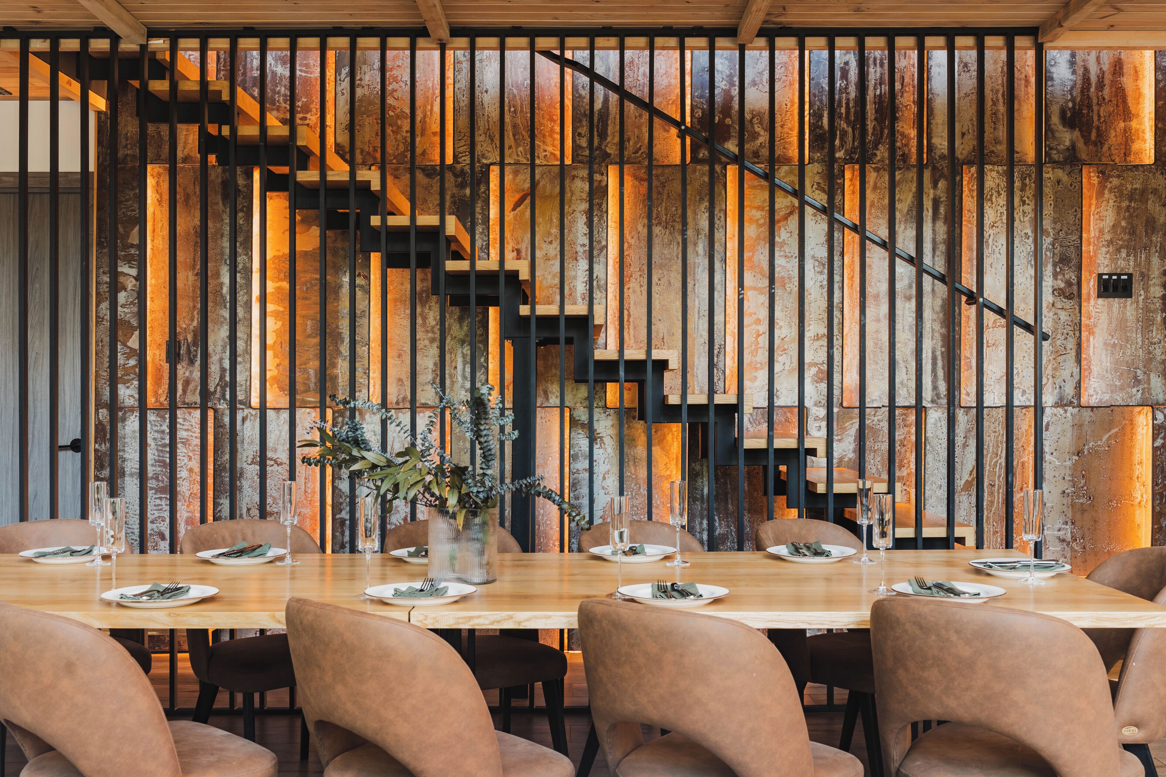 Ein Restaurant mit einem langen Holztisch, gepolsterten Stühlen und einer Wand mit vertikalen Holzleisten sowie einer Treppe.