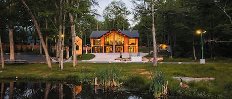 Une maison moderne avec un étang devant, entourée d’arbres.