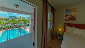 Ein Hotelzimmer mit Bett, Nachttisch mit Lampe, Fenster mit Vorhängen und Blick auf einen Swimmingpool sowie Gebäude im Freien.