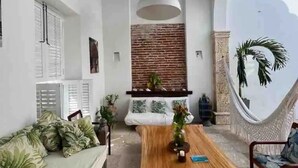 Ein Zimmer mit einem Holztisch, einer Couch mit grünen Kissen, einer Pendelleuchte und Blick durch ein großes, rundbogenförmiges Fenster.