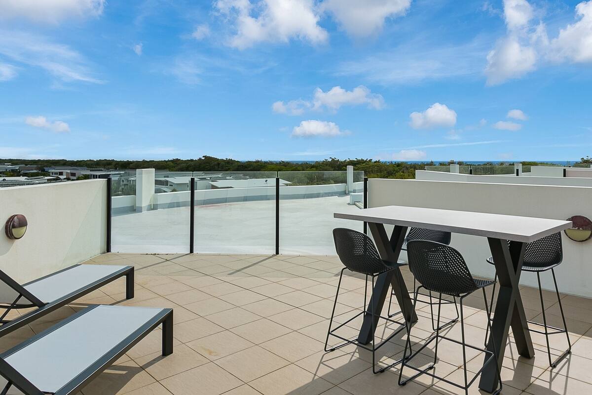 Une terrasse sur le toit, avec une table et des chaises, un banc et une balustrade en verre donnant sur un paysage.