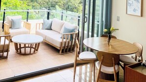 A modern living room with a sofa, chairs, a round table, and a balcony with a view of greenery and mountains. - Cotton Beach Resort (Casuarina)