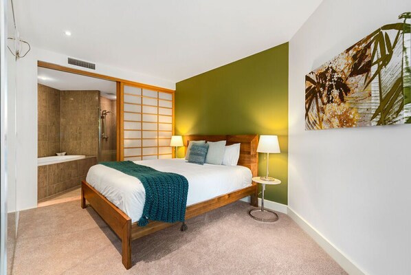 A bedroom with a wooden bed, a green accent wall, a bathroom with a shower, and a small round table with a lamp.