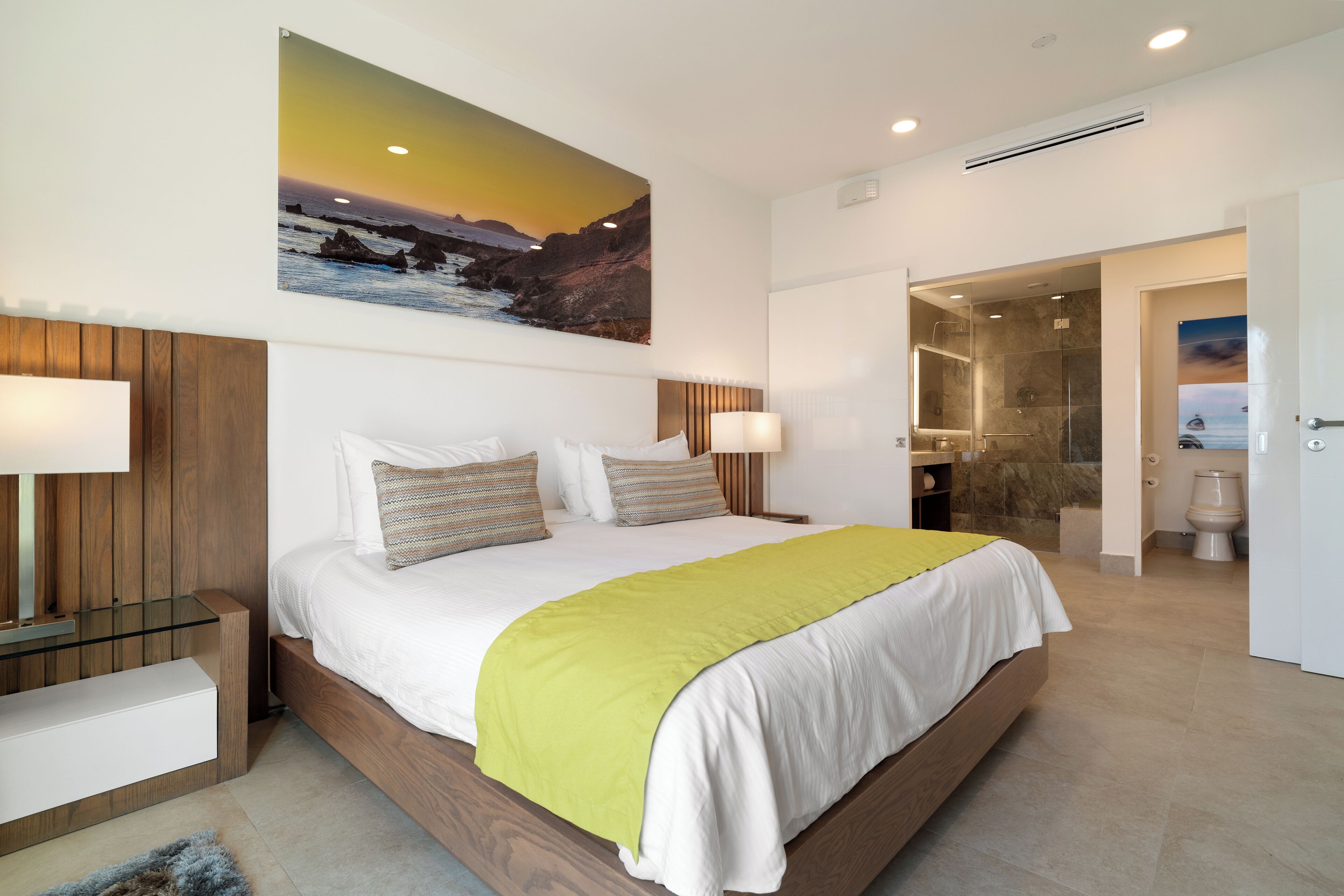 Una habitación de hotel moderna con una cama grande, cabecera de madera y un cuadro de una puesta de sol sobre el océano.
