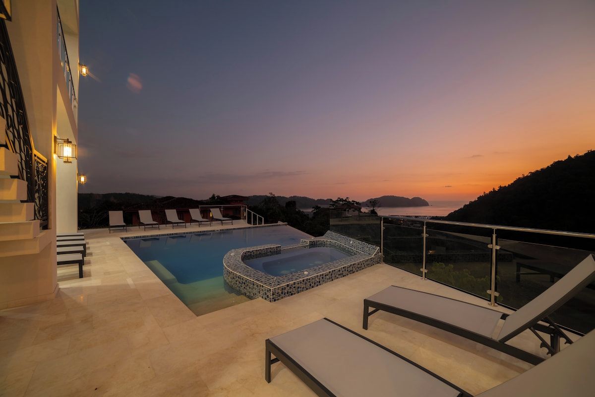 A rooftop pool with a hot tub, lounge chairs, and a view of the landscape.