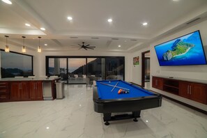 Una sala de estar moderna con mesa de billar, un televisor de pantalla plana grande y vistas a una isla tropical a través de amplios ventanales.