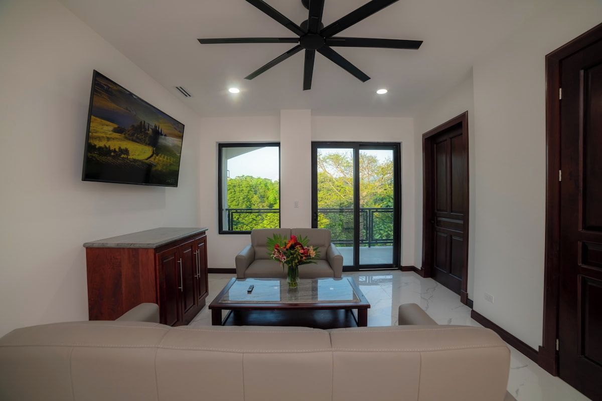 A modern living room with a ceiling fan, a sofa, a coffee table with a vase, and large windows with a view of greenery.