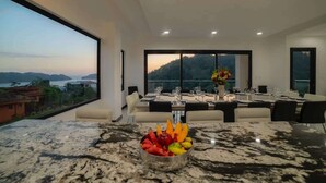 Un comedor moderno con un amplio ventanal que ofrece vistas a un paisaje pintoresco, una encimera de mármol con fruteros, y una mesa de comedor dispuesta para seis comensales.