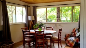 A dining area with a wooden table and chairs, a vase with flowers, and a view of greenery through large windows. - Idyllwylde: pet-friendly, renovated vintage  cabin, close to town and river (Warburton)