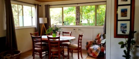 A dining area with a wooden table and chairs, a vase with flowers, and a view of greenery through large windows.