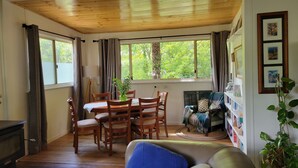 A cozy room with a wooden ceiling, a dining table with chairs, a sofa, a fireplace, and large windows with curtains. - Idyllwylde: pet-friendly, renovated vintage  cabin, close to town and river (Warburton)